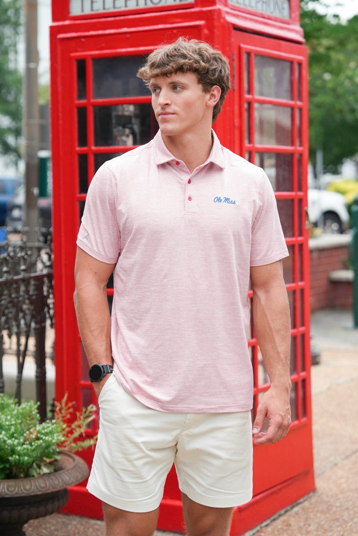 A man wearing a light pink polo shirt and off-white shorts stands in front of a classic red telephone booth. The polo shirt has a small blue embroidered logo on the left chest that reads 'Dr.