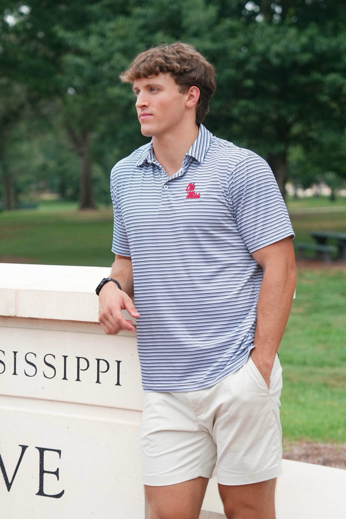 A young man wearing a blue and white striped polo shirt with a red logo on the chest, paired with off-white shorts, standing outdoors next to a stone wall with 'MISSISSIPPI' visible.