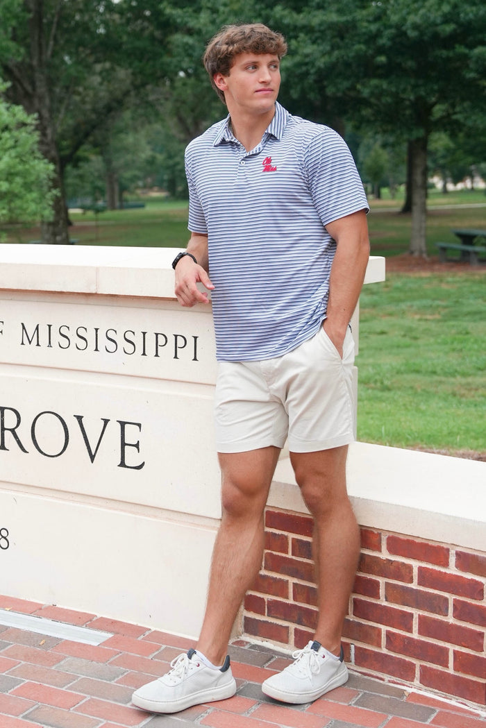 A man wearing a blue and white striped polo shirt with a red Mississippi logo, white shorts, and white sneakers, standing outdoors next to a brick and stone wall with 'MISSISSIPPI ROVE' text.