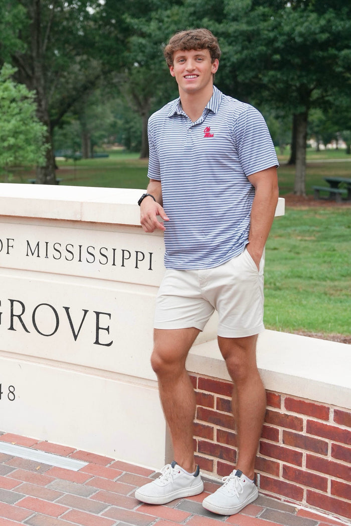 A young man stands outdoors leaning against a stone wall, wearing a blue and white striped polo shirt with a small red logo on the chest, khaki shorts, and white sneakers. The background shows a green park and a brick-and-stone sign that reads 'MISSISSIPPI GROVE'.