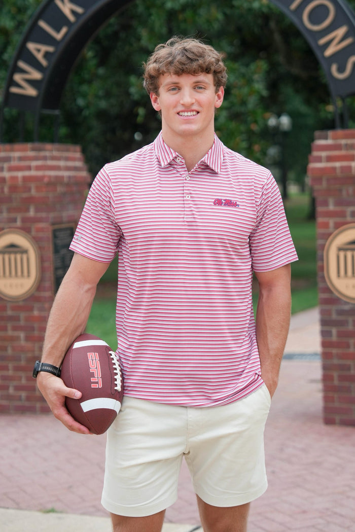 A young man stands outdoors holding a football, wearing a pink and white striped polo shirt with a small embroidered logo on the chest, paired with cream shorts.