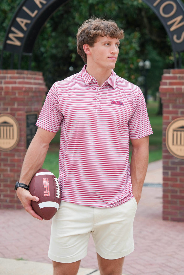 A man wearing a pink and white striped polo shirt with a small embroidered logo on the chest, paired with cream shorts, holding a football while standing in front of a brick archway.