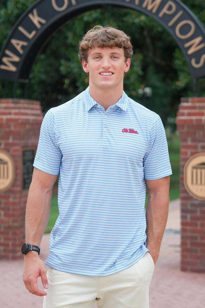 A young man smiling outdoors in front of a brick archway with a black sign that reads 'WALK OF CHAMPIONS'. He is wearing a light blue and white horizontally striped polo shirt with a red 'Old Miss' logo on the chest, paired with cream-colored pants.