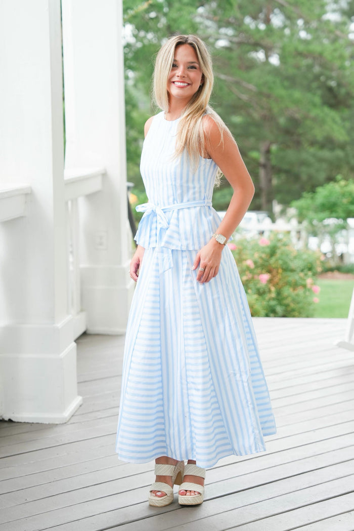 Woman in a light blue striped dress standing on a wooden deck with greenery in the background