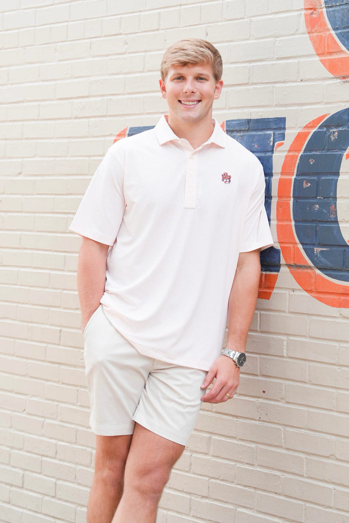 A smiling young man wearing a white short-sleeve polo shirt and matching white shorts, standing against a light brick wall with a painted mural partially visible behind him.