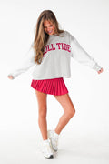 A woman modeling a light gray crewneck sweatshirt with 'COLUMBINE TIDE' printed in red across the chest, paired with a bright pink pleated mini skirt and white sneakers, against a plain white studio background.